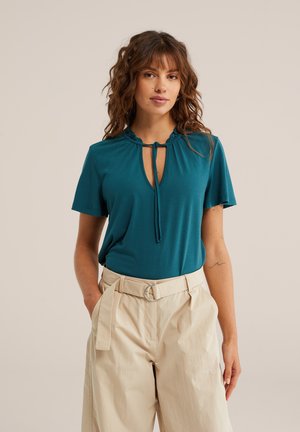 Žena s vlnitými hnedými vlasmi má na sebe tyrkysový top s krátkym rukávom a viazaním na výstrihu a béžové nohavice s vysokým pásom a opaskom, pózuje pred neutrálnym pozadím.