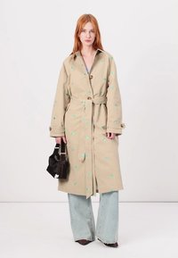 Beige trenchcoat met groene bloemenborduursel, knoopsluiting en strikriem; gedragen over lichtblauwe flared jeans; met een zwarte handtas.