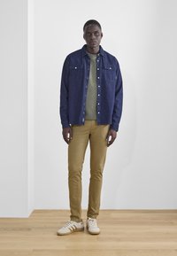 Camisa de mezclilla en azul oscuro, suéter verde oliva debajo, pantalones khaki slim-fit y zapatillas blancas con detalles en beige, de pie contra una pared blanca.