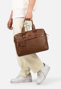 Malette en cuir brun avec doubles poignées supérieures, marquage en relief et étiquette bagages ; elle présente un design rectangulaire élégant avec fermeture éclair.