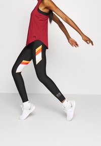 Bordeauxrode atletische tanktop met een mesh zak; zwarte leggings met oranje, witte en beige diagonale strepen; witte schoenen met grijze accenten.
