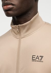 Μπεζ ζακέτα με φερμουάρ και ψηλό γιακά, κατασκευασμένη από υφασμάτινο ύφασμα με υφή, με μαύρο λογότυπο EA7 Emporio Armani στο στήθος.