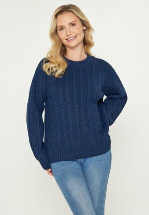 Femeie zâmbitoare cu păr ondulat blond, purtând un pulover din lână cu model de cablu în nuanță albastru închis și blugi deschiși, stând în fața unui fundal simplu.