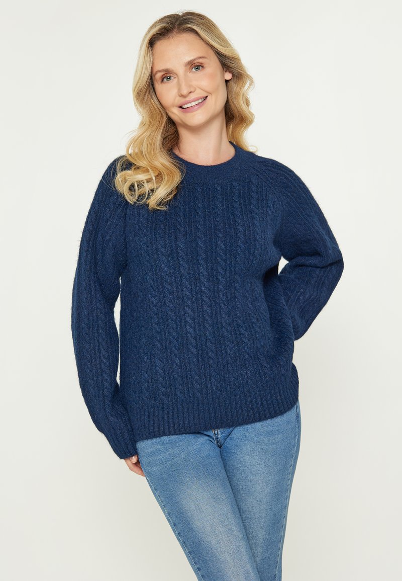 Donna sorridente con capelli biondi mossi, che indossa un maglione blu navy a trecce e jeans azzurri, in piedi davanti a uno sfondo semplice.