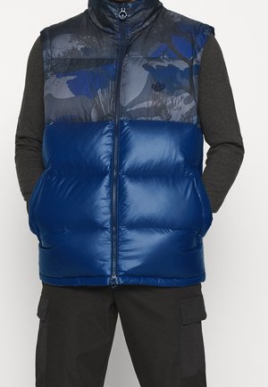 Veste matelassée bleue avec motif camouflage bleu foncé et gris sur la partie haute de la poitrine, portée sur des manches longues foncées et un pantalon cargo noir.
