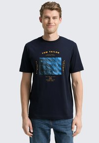 Granatowa koszulka z krótkim rękawem, wykonana z bawełny, z centralnym wzorem graficznym w postaci niebieskich spiral oraz pomarańczowych akcentów tekstowych.