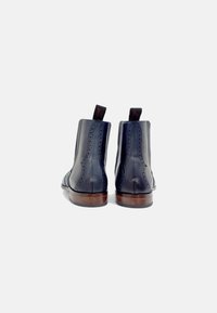Par de botas de cuero azul marino pulido hasta el tobillo con detalles de brogue y suelas de madera, vistas desde atrás sobre un fondo blanco.