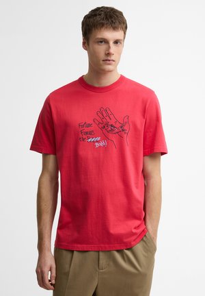 Młody mężczyzna w czerwonym T-shircie z grafiką dłoni i ryby oraz napisem „Fortune Favours the Bold!” stoi na białym tle.