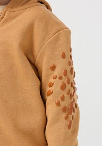 Felpa marrone con un design liscio e testurizzato, ornata da accenti lucidi in rilievo di diverse dimensioni sulla manica destra.