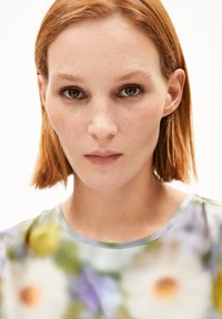 Bunt gemusterte Bluse mit rundem Ausschnitt. Der Stoff hat eine glatte Textur und zeigt Blüten in den Farben Grün, Lila und Gelb.