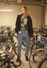 Sort vandtæt jakke, grafisk t-shirt, høj talje blå jeans, sorte snørebåndssko, omgivet af cykler i et parkeringsområde.
