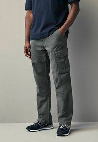 Graue Cargo-Hosen aus Baumwolle mit mehreren Seitentaschen, kombiniert mit navyblauen Laufschuhen mit weißen Akzenten und Schnürsenkeln.
