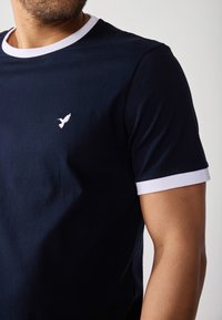 Námornícky modré tričko s bielym výstrihom a lemami rukávov. Obsahuje malé, vyšívané logo vtáka na hrudi. Jemná bavlnená tkanina.