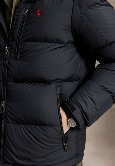 Black quilted jacket with red logo on chest, hand in zipped pocket, and wristwatch visible under sleeve cuff.