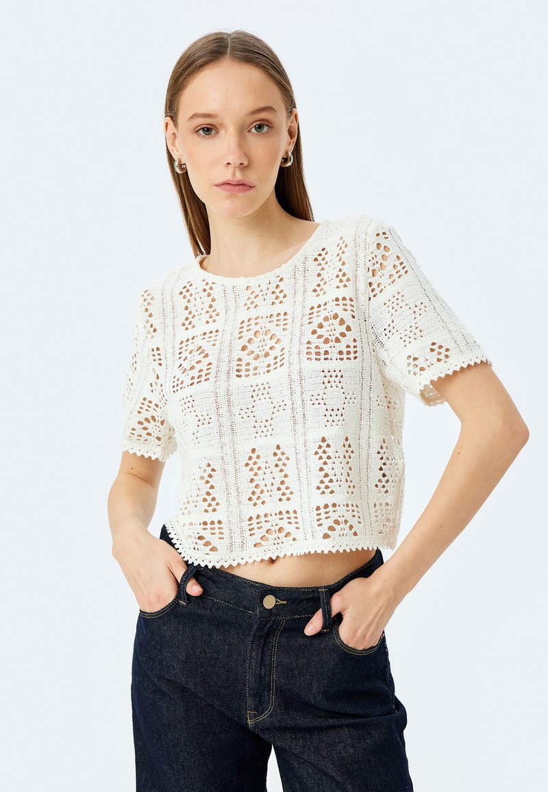 Top corto de croché blanco con mangas cortas, que presenta patrones intrincados y un dobladillo en forma de ondas. Combinado con jeans de mezclilla oscura.