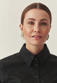 Fekete galléros ing gombokkal, sima textúrával. A modellen nagy, ezüstszínű fülbevalók, lágy semleges háttér.