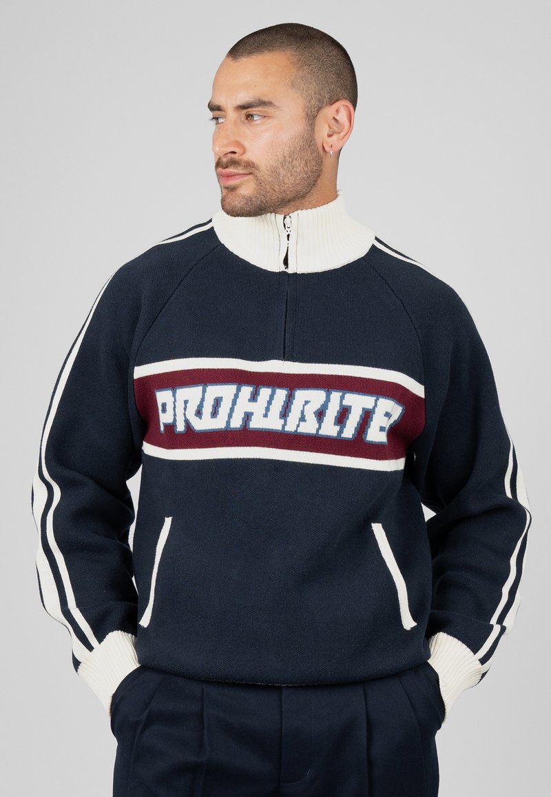 Homme portant un pull bleu marine à rayures blanches avec le mot "PROHBLITE" en grandes lettres blanches et bleues sur la poitrine.