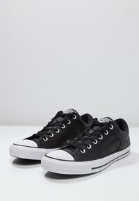 Černé kožené tenisky s bílými gumovými podrážkami, kulatou špičkou, černými tkaničkami a klasickou nášivkou loga na jazyku. Minimalistický design s černými pruhy.