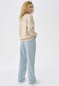 Beige gebreide trui met getextureerde accenten, gecombineerd met lichtblauwe geruite wijde broek en witte sneakers. Model staat tegen een neutrale achtergrond.