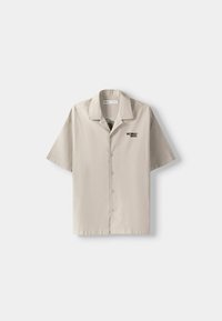 Camisa de manga corta con botones en algodón beige claro, con un logo negro "NOMAD" en el pecho izquierdo y diseño de cuello clásico.