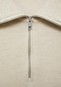 Beige lynet pullover med et tekstureret stof. Har en metal lynlås og en kravedesign. Mønstret strikoverflade er synlig.