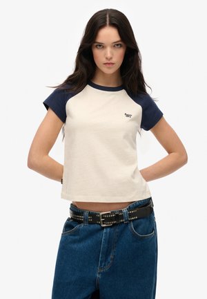 Jeune femme avec de longs cheveux bruns portant un t-shirt à manches raglan beige et marine et un jean bleu avec une ceinture noire cloutée.