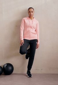 Roza zip-up kapuca v kombinaciji s črnimi trenirka hlačami in črnimi športnimi čevlji. Ima sprednjo kenguru žep in logotip na kapuci.