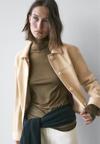 Lysebeige jakke med runde knapper, båret over en grøn rullekrave. Et mørkt strikket tørklæde er bundet om taljen og skaber et lagdelt udseende.