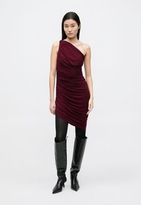 Vīnsarkanā vienas pleca kleita ar savilktu tekstūru, pieguļošs siluets, kombinēta ar melniem ceļgalu augstiem zābakiem un necaurspīdīgām zeķbiksēm.