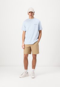 Tricou din bumbac de culoare albastru deschis, cu mâneci scurte, pantaloni șort bej și adidași albi. Îmbrăcămintea include o șapcă bej, cu detalii de text minime.