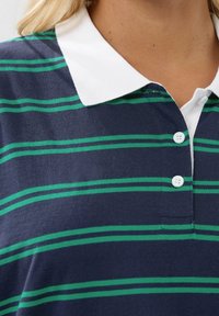 Marinblå polo-shirt med vit krage och gröna ränder, med tre knappar på knäppningen och en mjuk bomullstextur.