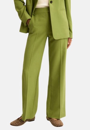 Donna con pantaloni a gamba larga verde oliva e blazer abbinato, scarpe basse marroni a pois, in piedi con una mano in tasca.