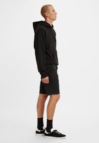 Svart hettegenser med kengurulomme, kombinert med mørke shorts og svarte joggesko. Teksturert stoff, avslappet passform og minimalistisk design.