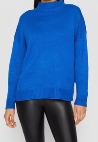 Pull bleu tricoté avec un col montant, des poignets et un ourlet côtelés. Porté avec un pantalon noir ajusté. La texture semble douce et confortable.