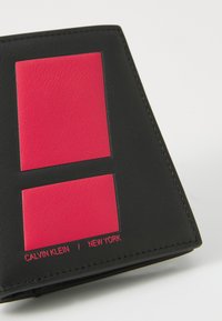 Portofel din piele neagră cu două accente roșii rectangulare. Textul de brand în roșu, "CALVIN KLEIN / NEW YORK," aproape de marginea de jos.