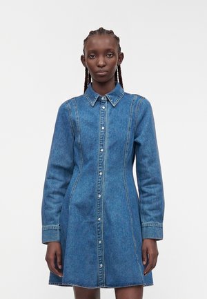 Femme aux cheveux tressés portant une robe en jean bleu à manches longues, boutonnée, avec un col, debout devant un fond blanc uni.