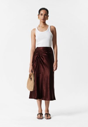 Bronze satin skirt with a ruched detail at the waist, paired with a white tank top, straw bag, and brown flat sandals.