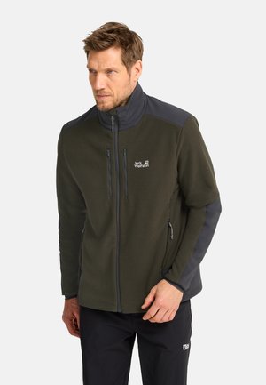 Man draagt een groen-zwart Jack Wolfskin jas met rits en hoge kraag en zwarte broek, staand tegen een effen achtergrond.