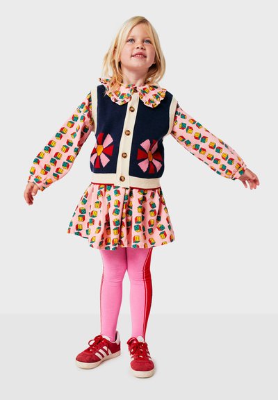 Chaleco de punto con diseños florales, cierre frontal de botones, sobre un vestido estampado en rosa. Leggings rosas con rayas laterales y zapatillas rojas.