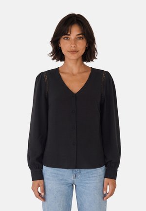 Femme aux cheveux courts et foncés portant un chemisier noir à manches longues boutonné et un jean bleu clair sur un fond clair uni.