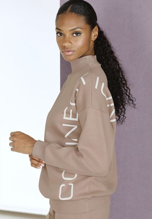 Light brown sweatshirt with high neck, featuring white text design on sleeves. Soft texture, relaxed fit, ribbed cuff, and hem.
