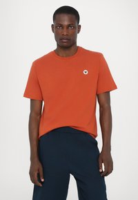 Oranžs kokvilnas T-krekls ar īsām piedurknēm, apaļu dekoltē un mazu, baltu, apaļu logotipu kreisajā krūtī. Saskaņots ar tumši ziliem biksēm.