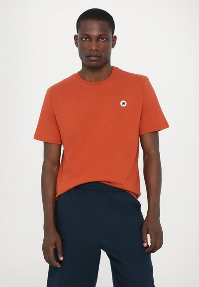 Orange cotton t-shirt with short sleeves, round neckline, and a small circular white logo on the left chest. Paired with navy pants.