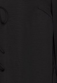 Haut noir en tissu léger et texturé. Présente un ourlet incurvé subtil et une fente sur le côté, avec des liens attachés à l'avant.