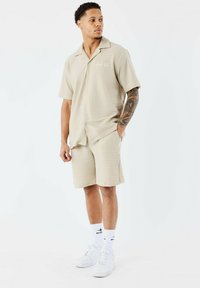 Camisa beige de manga corta con diseño de cuello, que presenta un sutil patrón texturizado. Combinada con pantalones cortos a juego, zapatillas blancas y calcetines.