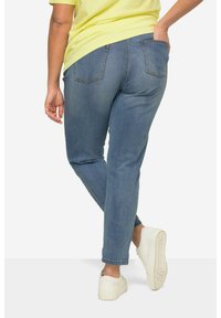 Jeans de mezclilla azul con pierna recta, bolsillos traseros y una textura suave, combinados con zapatillas blancas y una camisa amarilla.