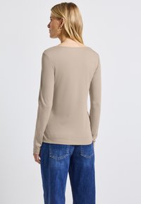 Pitkähihaisessa beige puserossa on avara kaula-aukko, pehmeä kangas ja tiukka istuvuus. Puseroa on yhdistetty korkeavyötäröisiin sinisiin farkkuihin, joissa on takataskut.