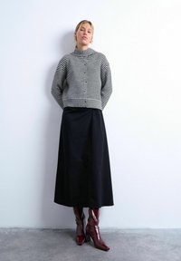Femme debout contre un mur blanc, portant un pull boutonné rayé noir et blanc, une jupe midi noire et des bottes bordeaux brillantes.