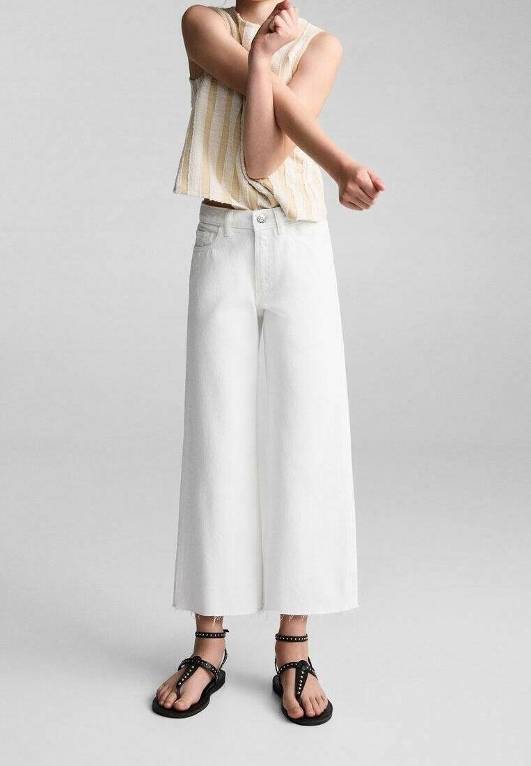 Personne portant un pantalon large blanc raccourci, un haut rayé sans manches et des sandales noires cloutées sur un fond clair uni.