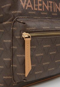 Bolsa de couro texturizado castanho com detalhes em metal dourado, um bolso com fecho e padrão de logótipos repetidos na superfície.